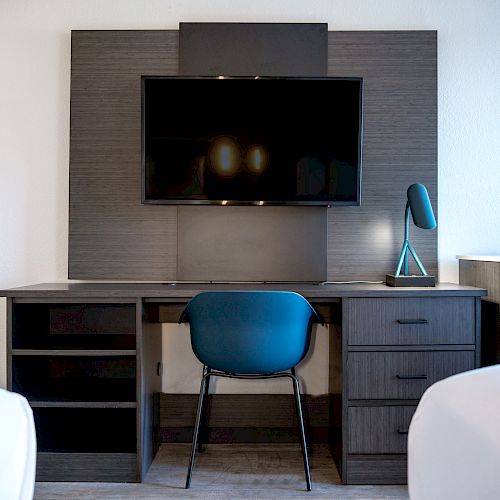 A modern desk setup features a mounted TV, blue chair, desk lamp, and drawers, with two beds visible on either side ending the sentence.