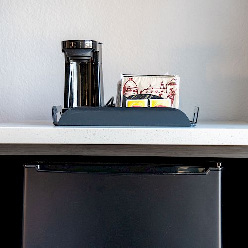 The image shows a small mini-fridge with a white countertop. A tray with a coffee maker, coffee pods, and cups is placed on the countertop.