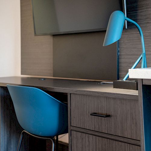 A modern workspace with a desk, blue chair, blue desk lamp, and a wall-mounted screen. The desk has shelves and drawers for storage, ending the sentence.