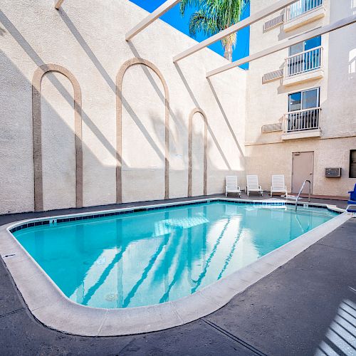 The image shows a clear, rectangular swimming pool in a sunny, concrete patio area, surrounded by white walls and several lounge chairs.