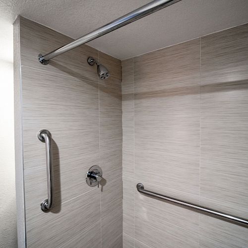 This image shows a shower area with beige tiles, a showerhead, and two stainless steel grab bars for safety.