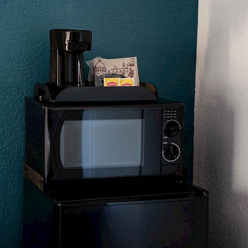 The image shows a microwave on top of a mini-fridge, with a coffee maker and some items, possibly tea bags, placed on top of the microwave inside a room.