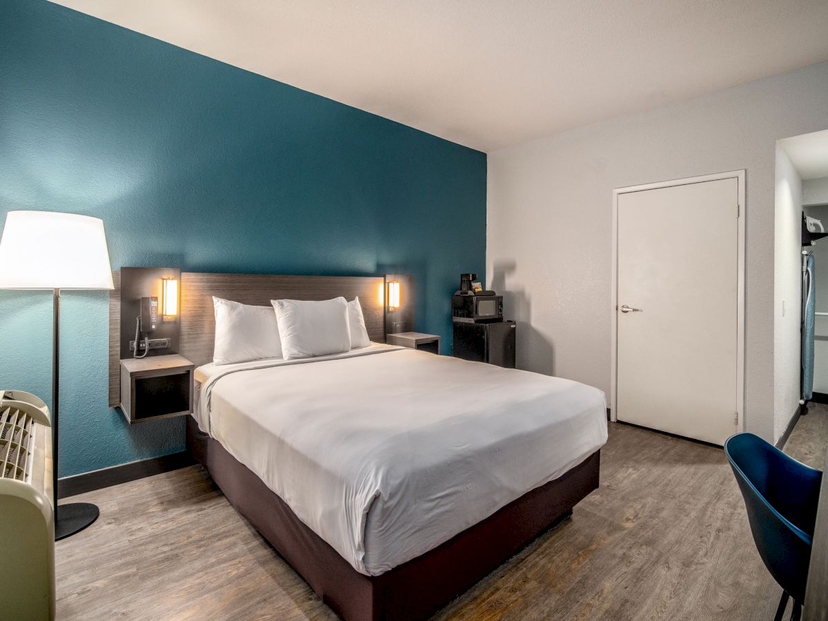 A modern hotel room with a double bed, teal accent wall, bedside lamps, a desk, chair, lamp, air conditioner, and wooden flooring.