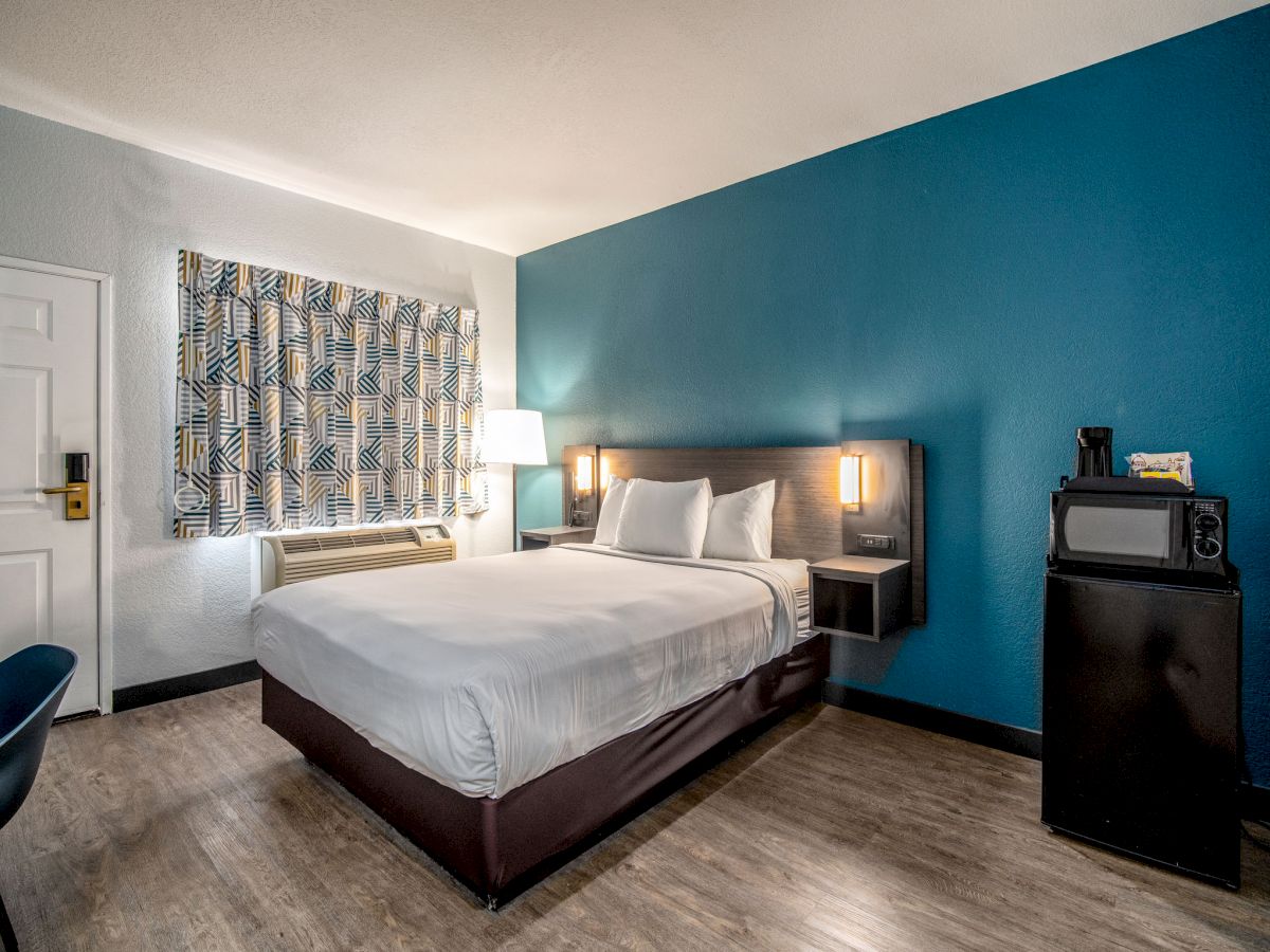 A modern hotel room with a bed, blue accent wall, bedside lamps, a window with curtains, a mini-fridge, a microwave, and a desk with a chair.