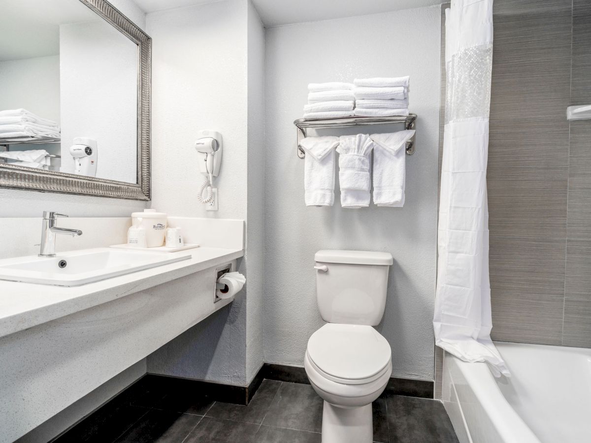 A clean bathroom with a toilet, sink, mirror, hairdryer, and a rack of neatly folded towels above the toilet, adjacent to a bathtub with a shower curtain.