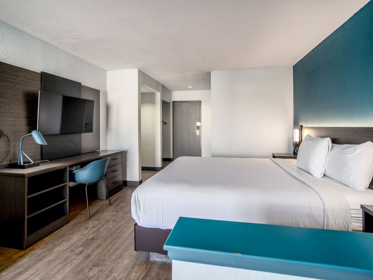 Modern hotel room with a large bed, desk, chair, flat-screen TV, and blue accents. Room is clean, with wood floors and ample natural light.