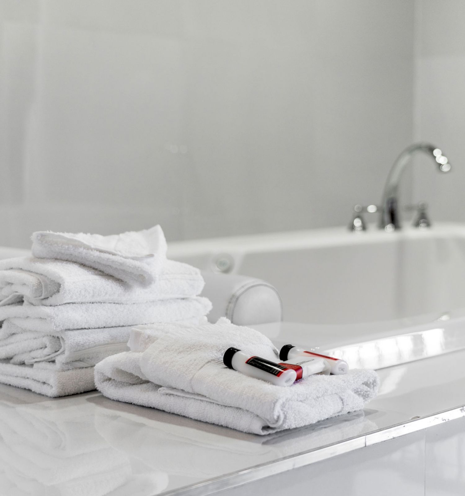 A clean, white bathroom with a pile of folded towels and two toothbrushes placed on the counter near a bathtub with a sleek faucet.