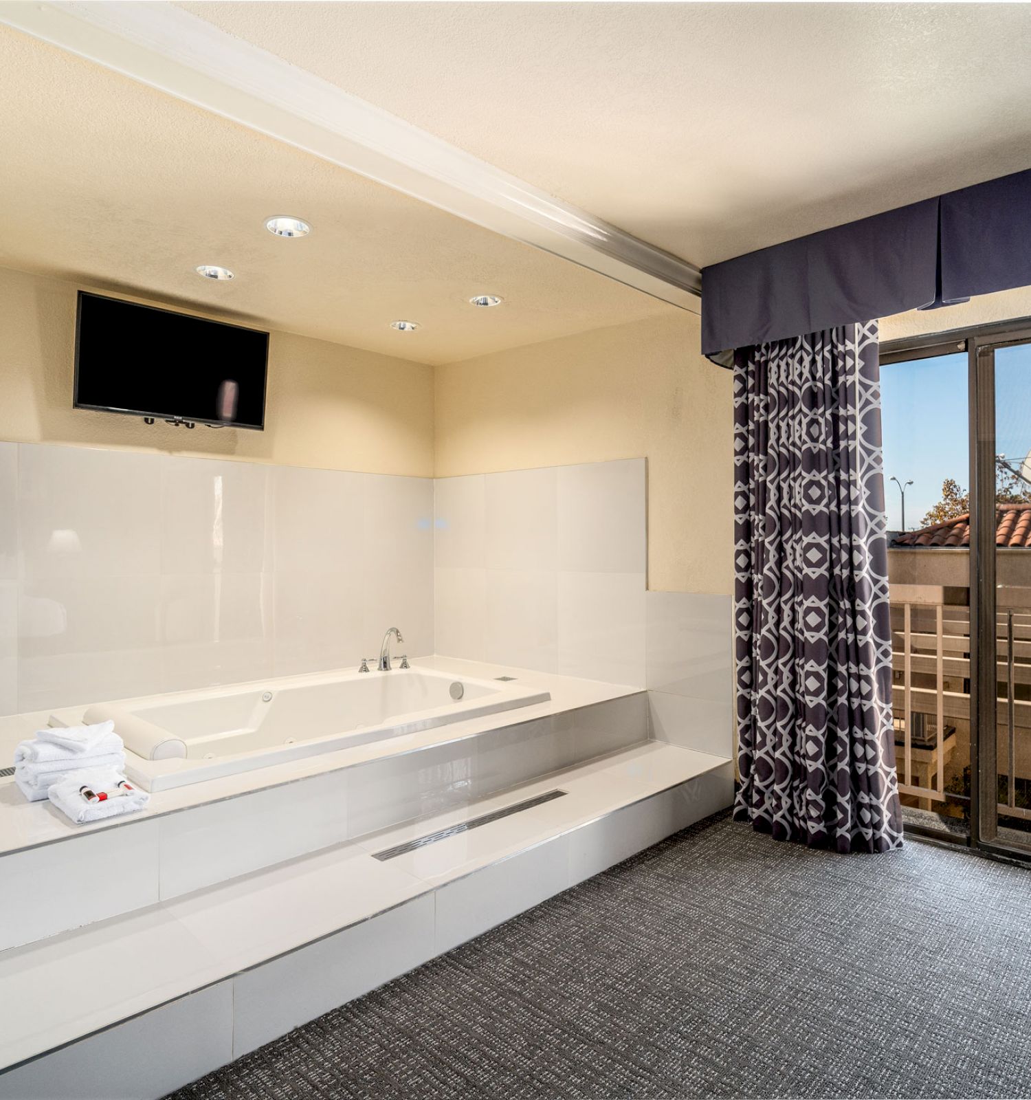 The image shows a modern room with a large bathtub, a TV above it, and a window with patterned curtains leading to a small balcony with a view.