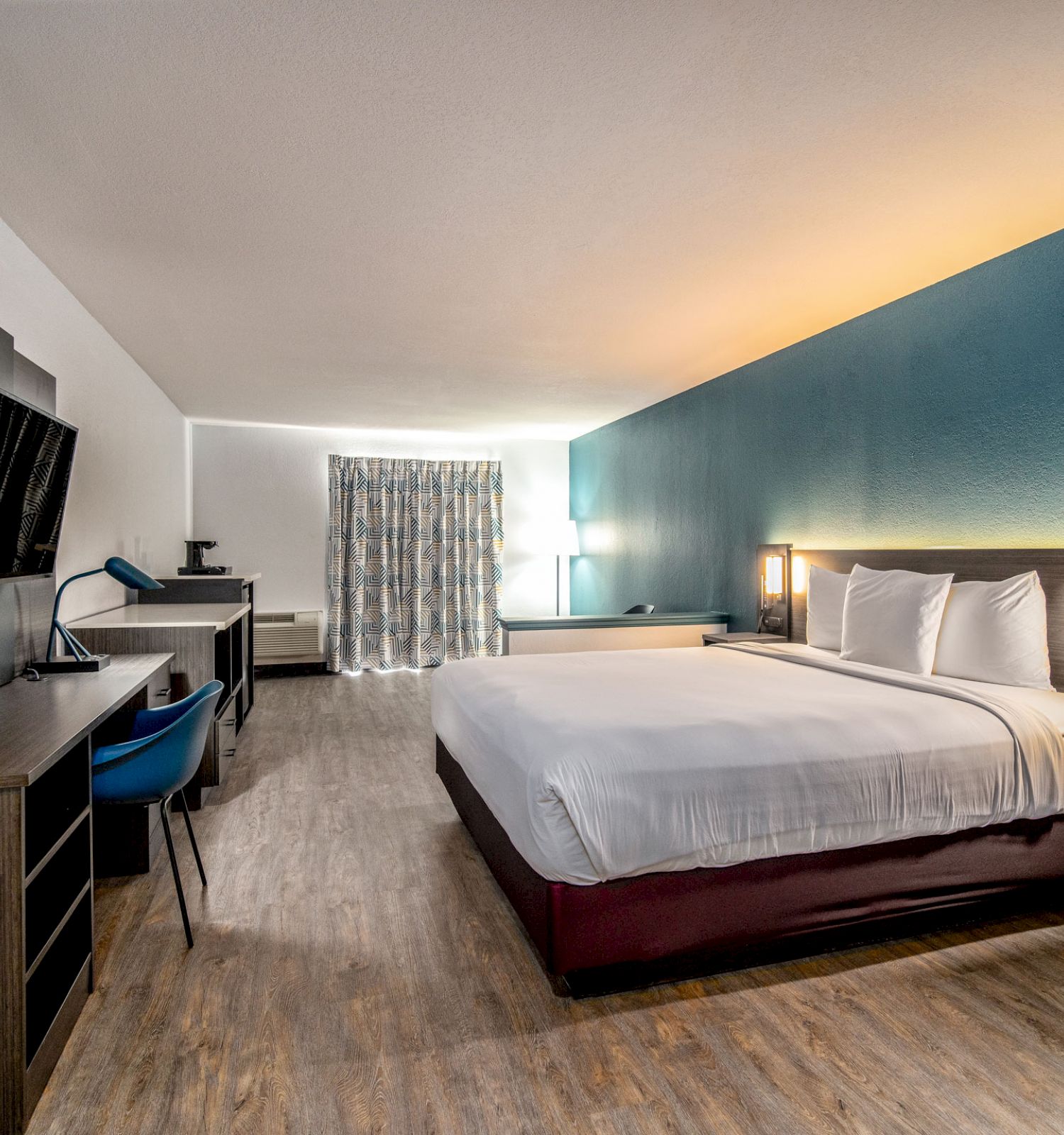 A modern hotel room with a king-sized bed, TV, desk, chair, and a blue accent wall, along with wooden flooring and a window with curtains.