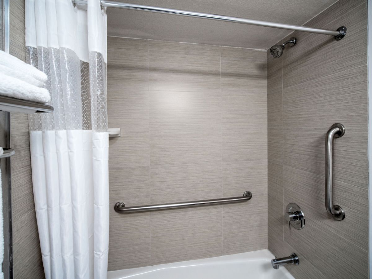 The image shows a clean, modern bathroom shower area with a shower curtain, grab bars, a showerhead, and a towel rack with neatly stacked towels.