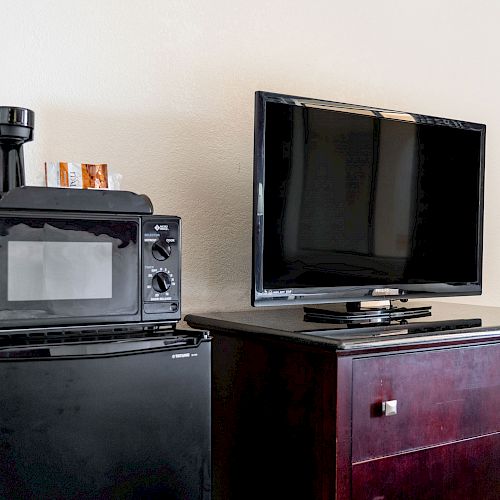 The image shows a small setup with a black microwave, coffee maker, and mini-fridge next to a dark wooden dresser with a flat-screen TV on top.