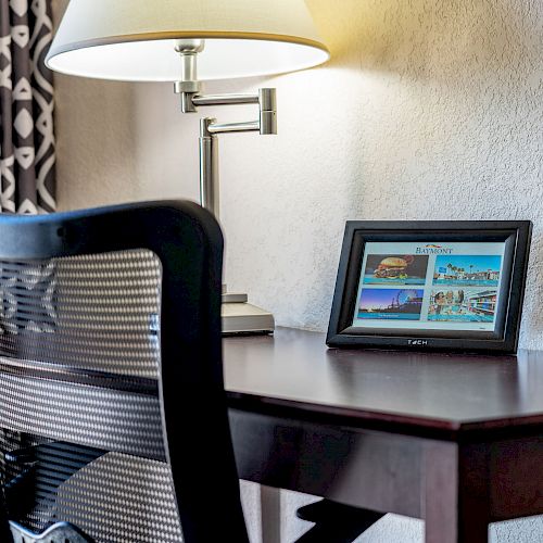 A wooden desk with a black chair, a lamp, and a framed photo displaying various colorful images.