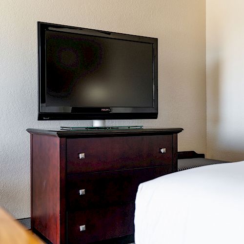 A flat-screen TV sits on a wooden dresser with drawers. The corner of a bed and a microwave in a small kitchen nook are also visible.