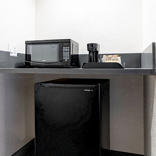 The image shows a small kitchenette area with a black mini-fridge, a microwave, and a coffee maker placed on a countertop.