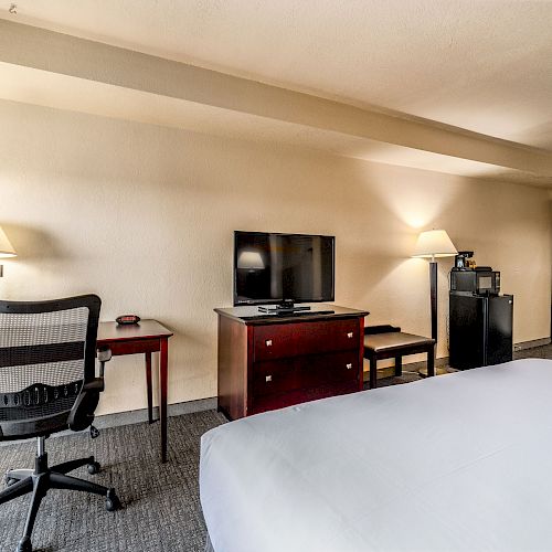 A hotel room features a bed, desk, chair, TV, lamps, and a mini fridge with a microwave on top.