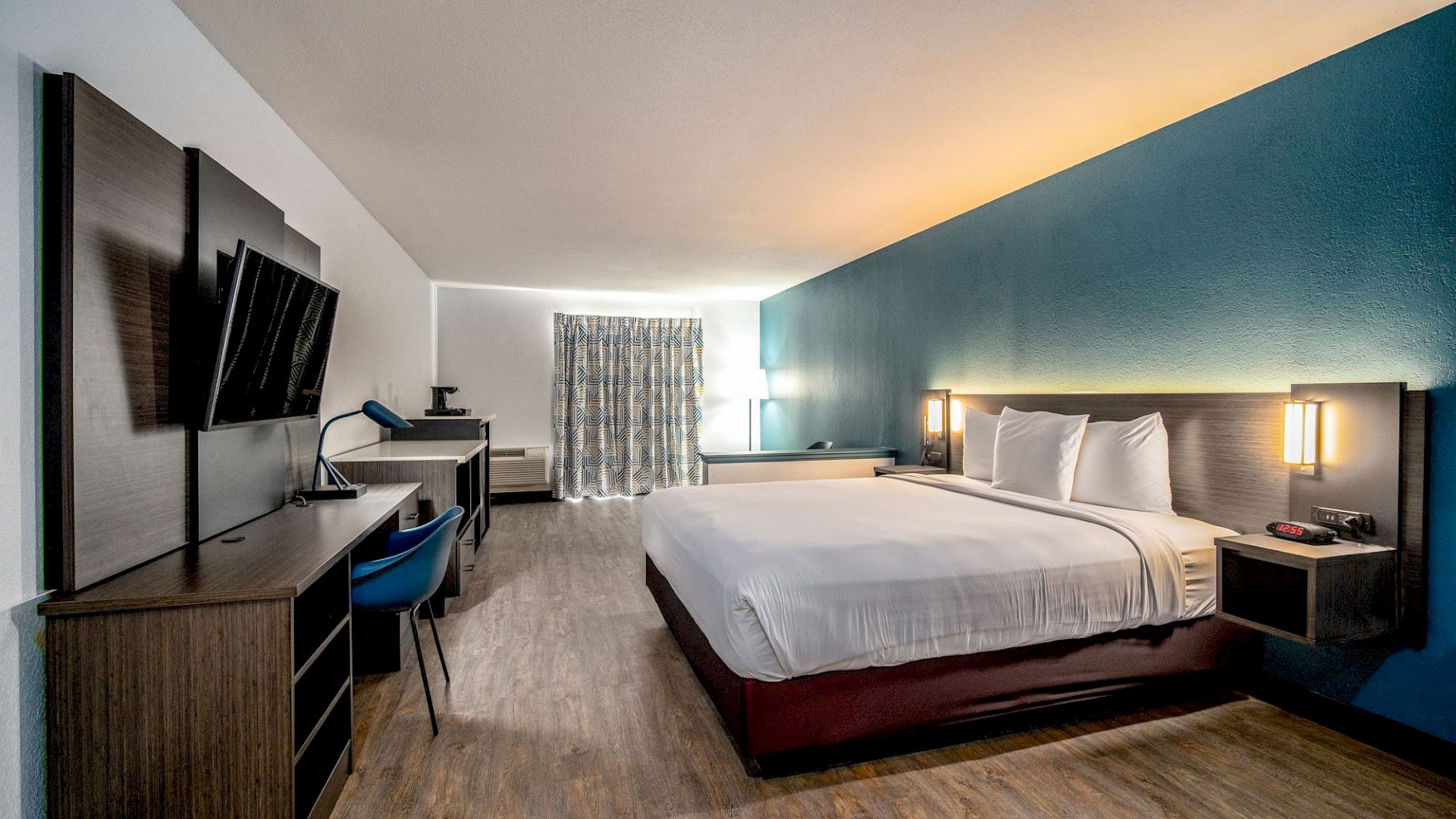 A modern hotel room with a large bed, wall-mounted TV, desk, chair, and a window with curtains. The decor is simple and clean.