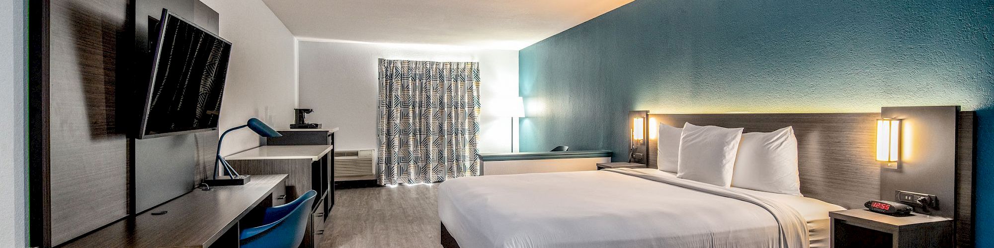 A modern hotel room with a large bed, wall-mounted TV, desk, chair, and a window with curtains. The decor is simple and clean.