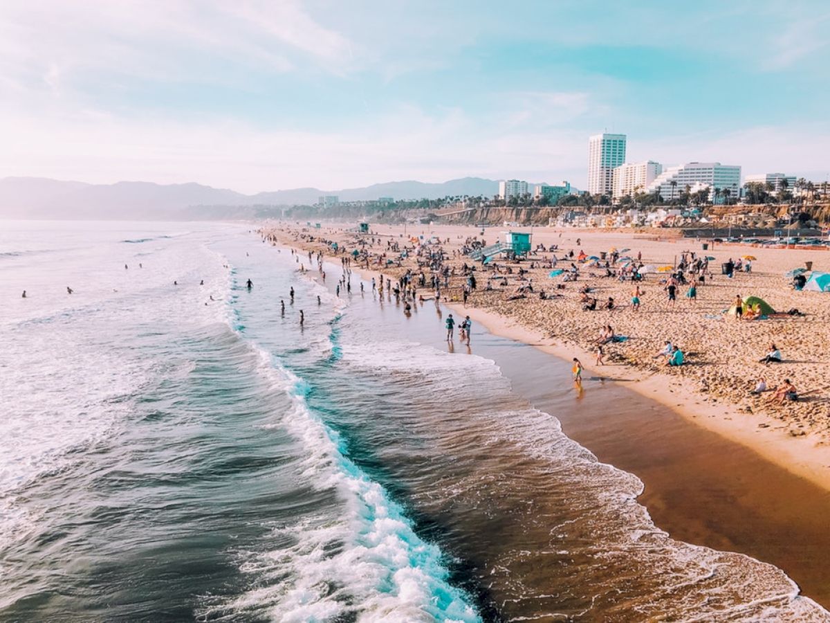 The image shows a crowded beach with people enjoying the sand and sea, with buildings and mountains visible in the background under a clear sky.