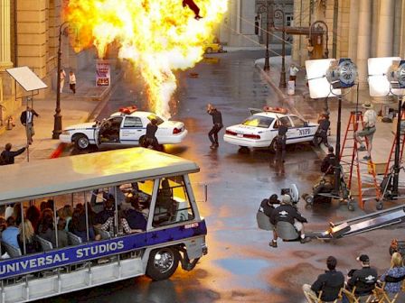 A film set with an explosion, police cars, a tour bus, and crew members filming the scene with lighting and camera equipment.