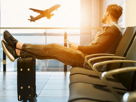 A man is sitting in an airport lounge with his legs on a suitcase, watching a plane take off through the window as light shines through.