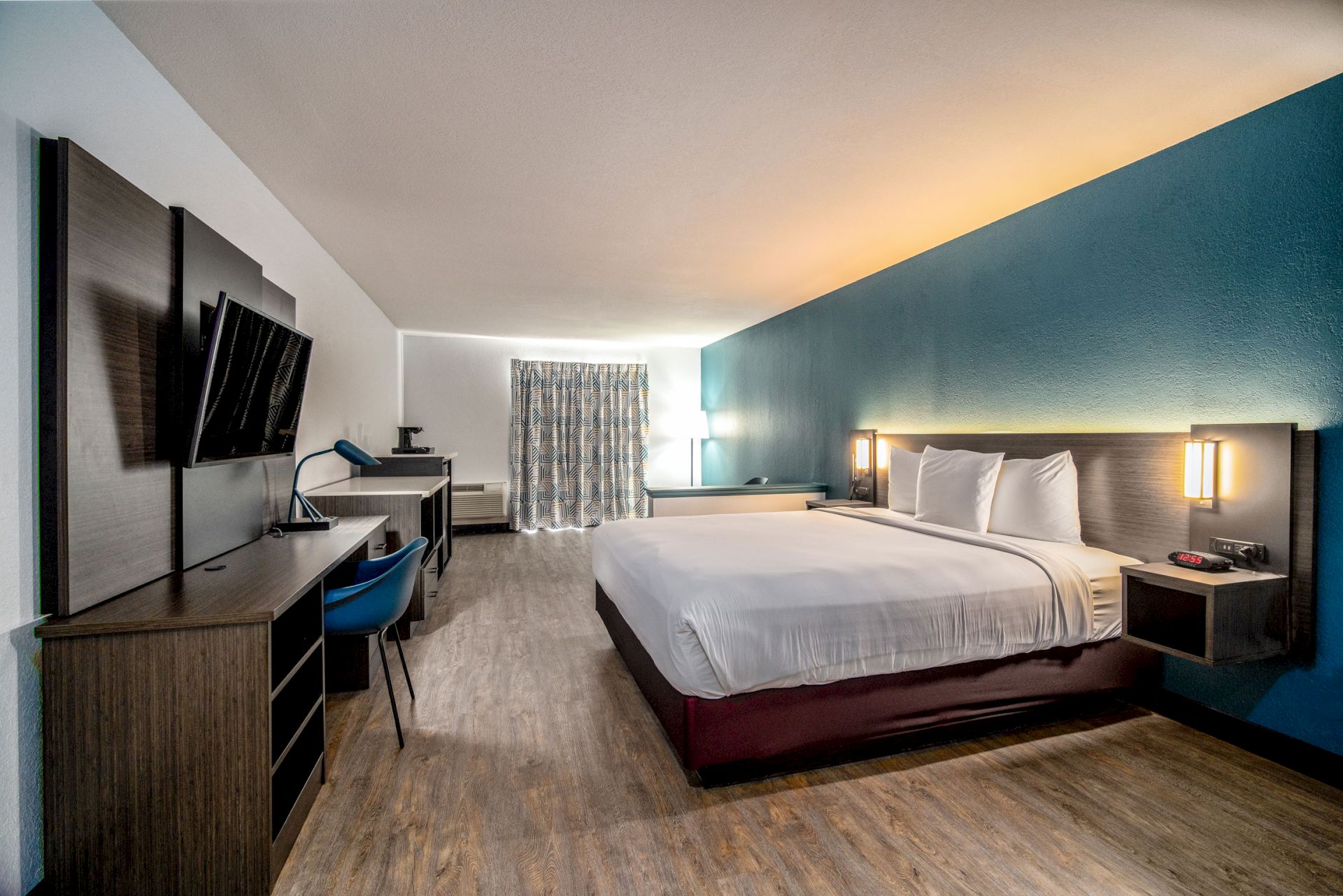 A hotel room with a king-size bed, mounted TV, desk, chair, and modern decor. The room has wooden flooring and ambient lighting.