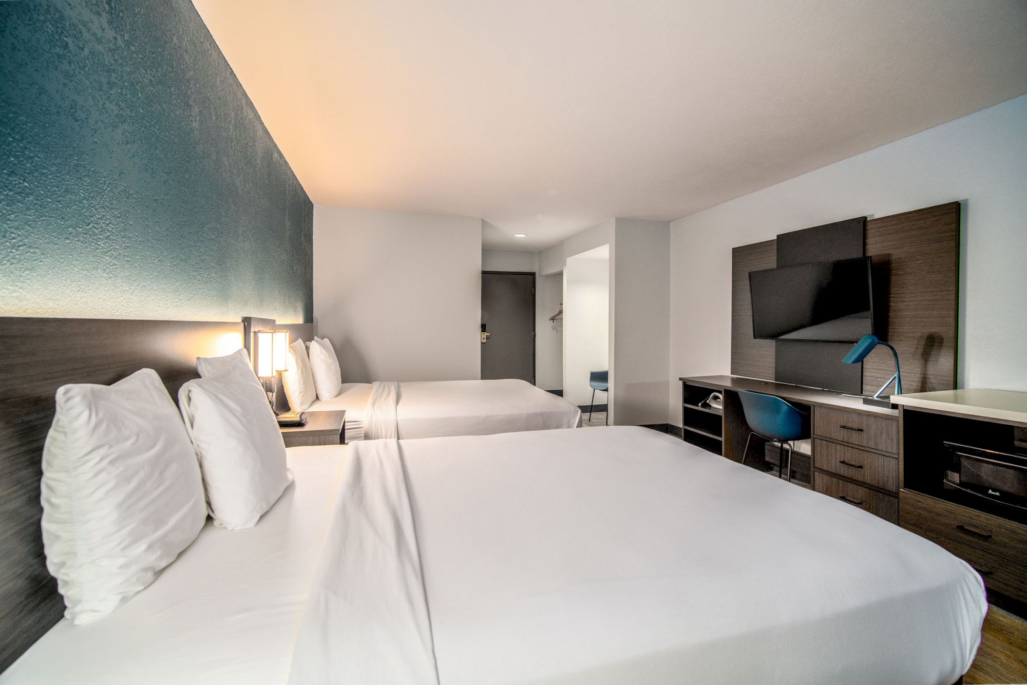 A modern hotel room with two double beds, a flat-screen TV, desk, chair, and a lamp, featuring a serene blue wall accent and minimalist decor.